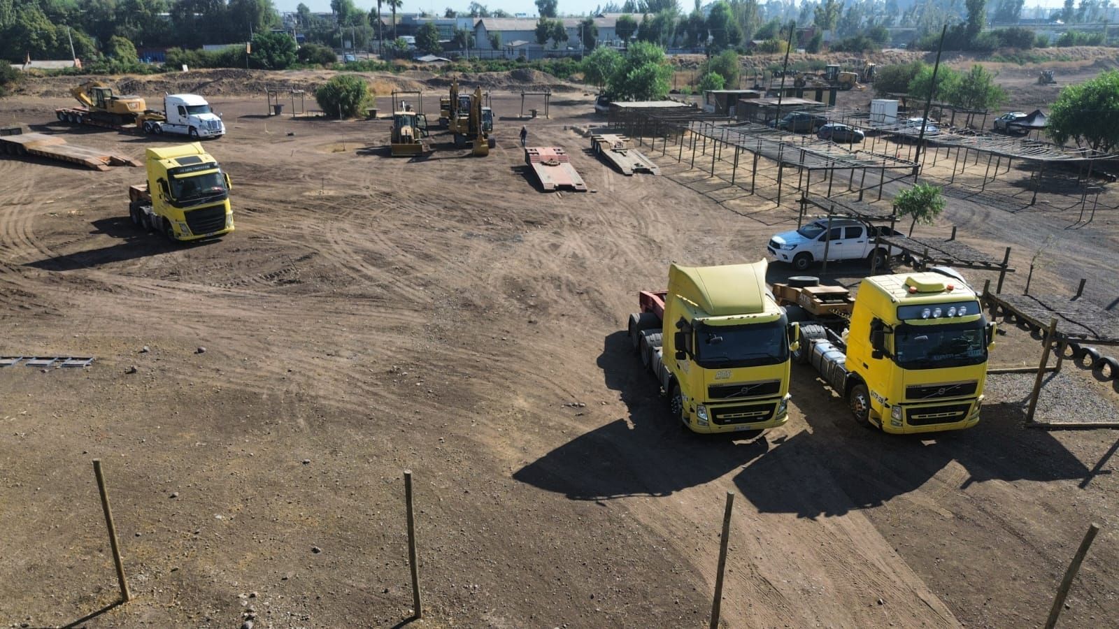 Tres camiones semirremolque amarillos estacionados en un lote de tierra con equipos de construcción