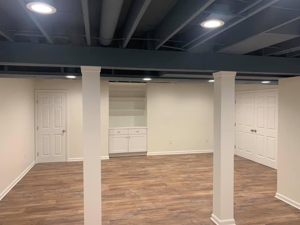 Finished basement with hardwood floor, white columns, built-in shelving, recessed lighting, and two white doors.