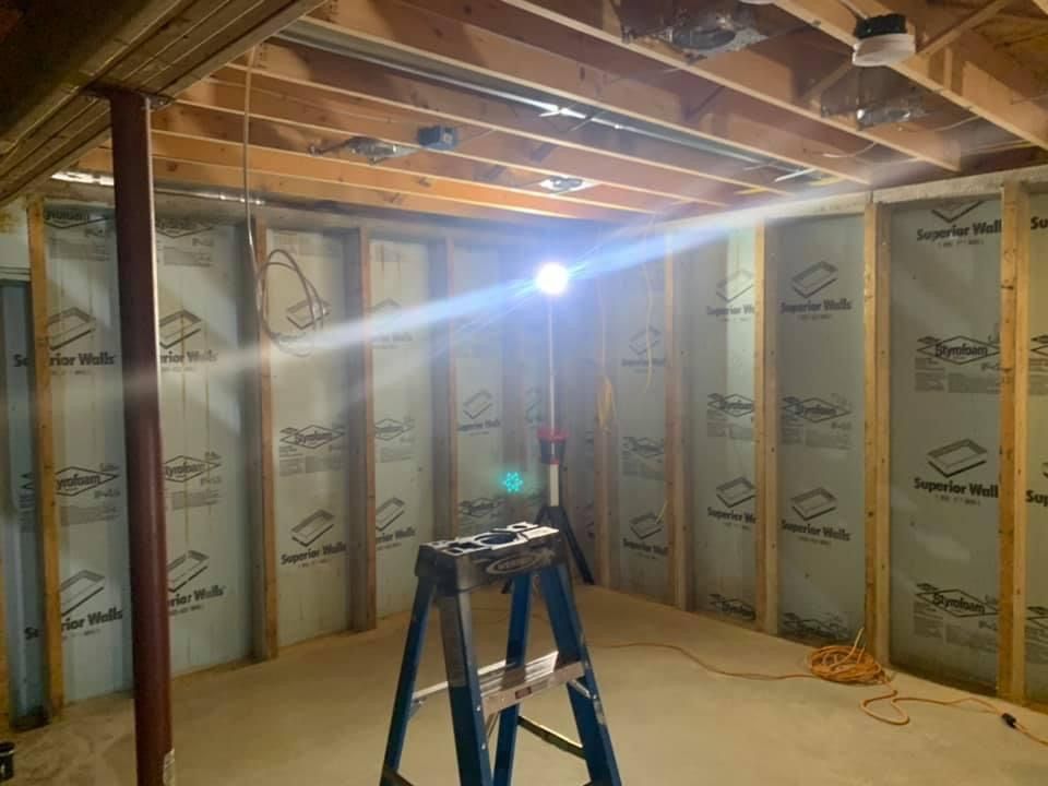 Interior view of a room under construction, lit by a work light. Studs and insulation are visible; a stepladder is in the foreground.