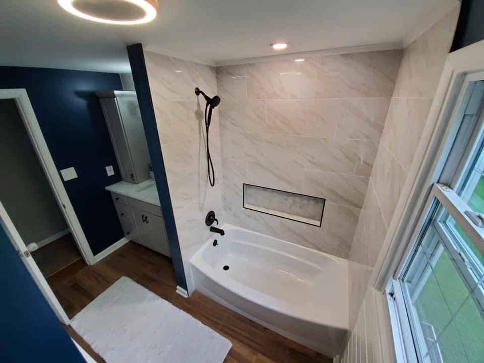 Bathroom with white tub, marble-look tile shower, dark blue walls, and wood floor.