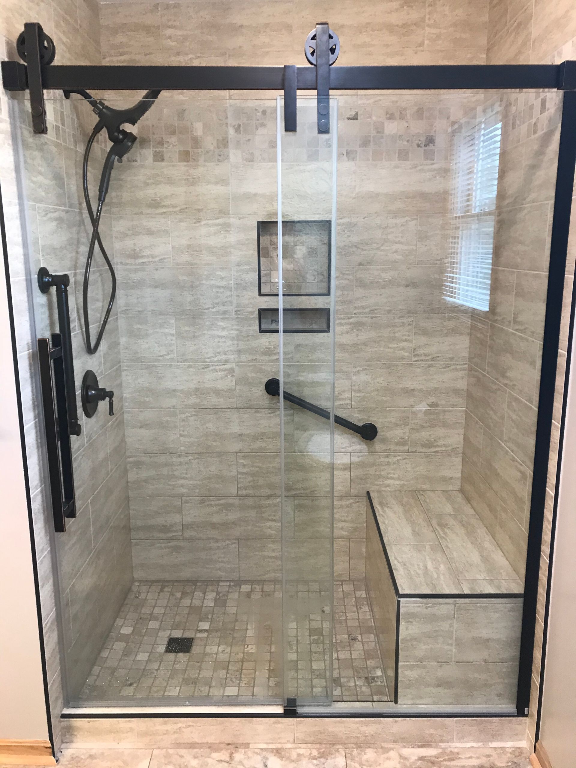 Accessible shower with sliding glass door, grab bar, and bench, tiled in beige.