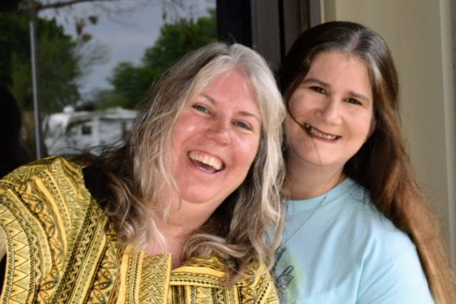 Two smiling women, one in yellow, one in blue, standing outside a building.