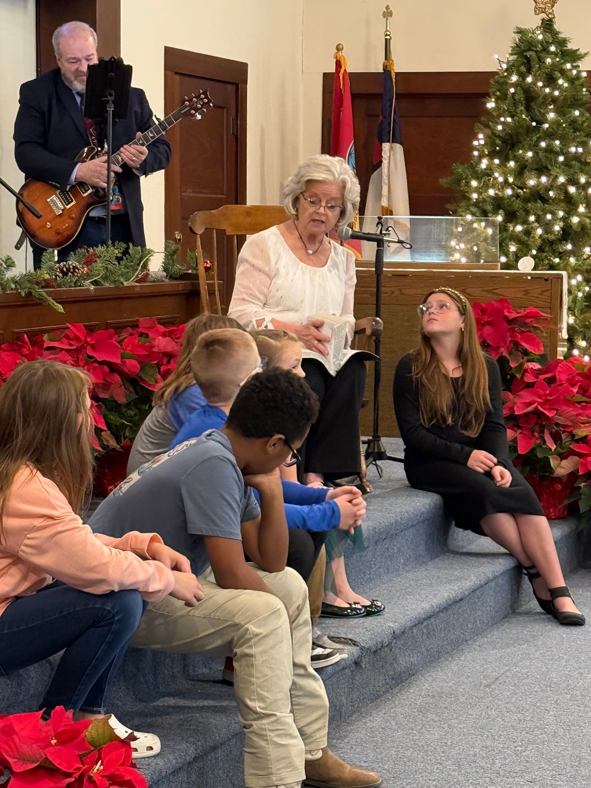 People singing and playing music on a stage decorated with poinsettias and a Christmas tree.