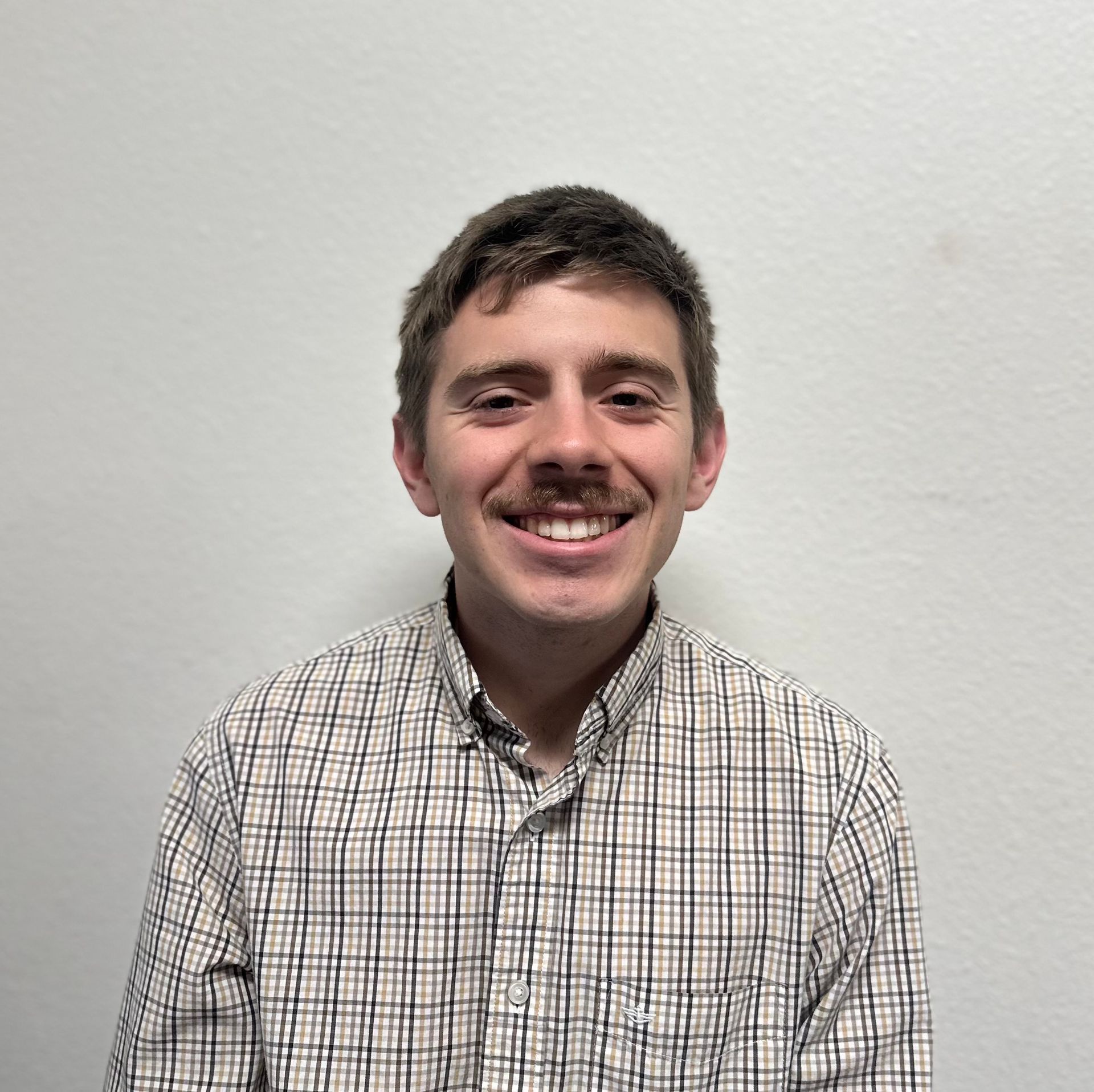 Man with mustache smiles, wearing plaid shirt, against a white wall.