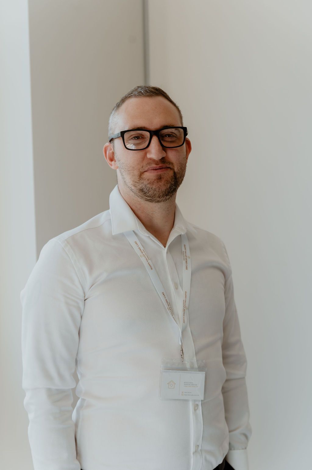 Man in a white shirt and glasses standing indoors against a plain wall, wearing a lanyard badge.