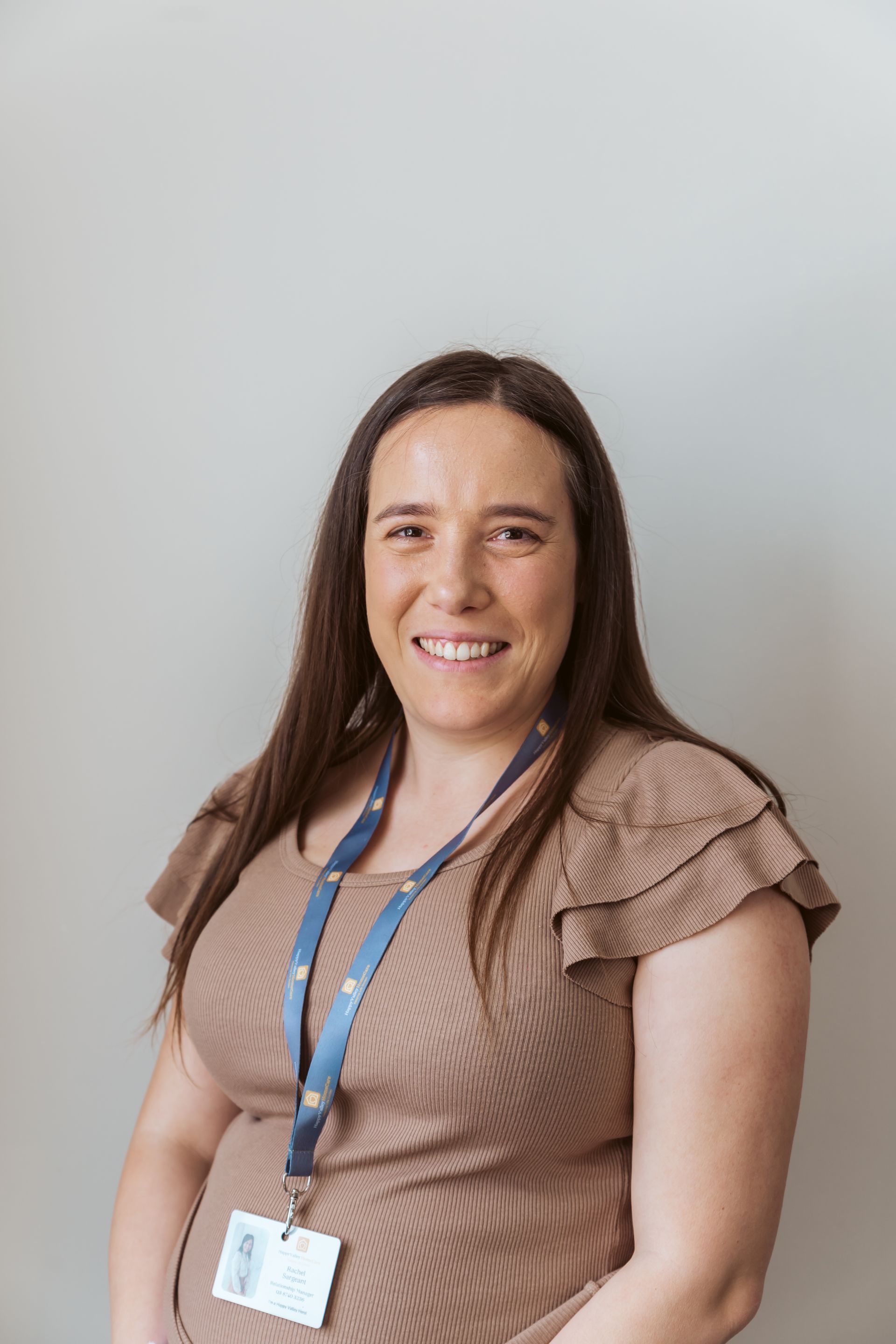 Smiling woman in a tan top with a blue lanyard, seated against a light gray background