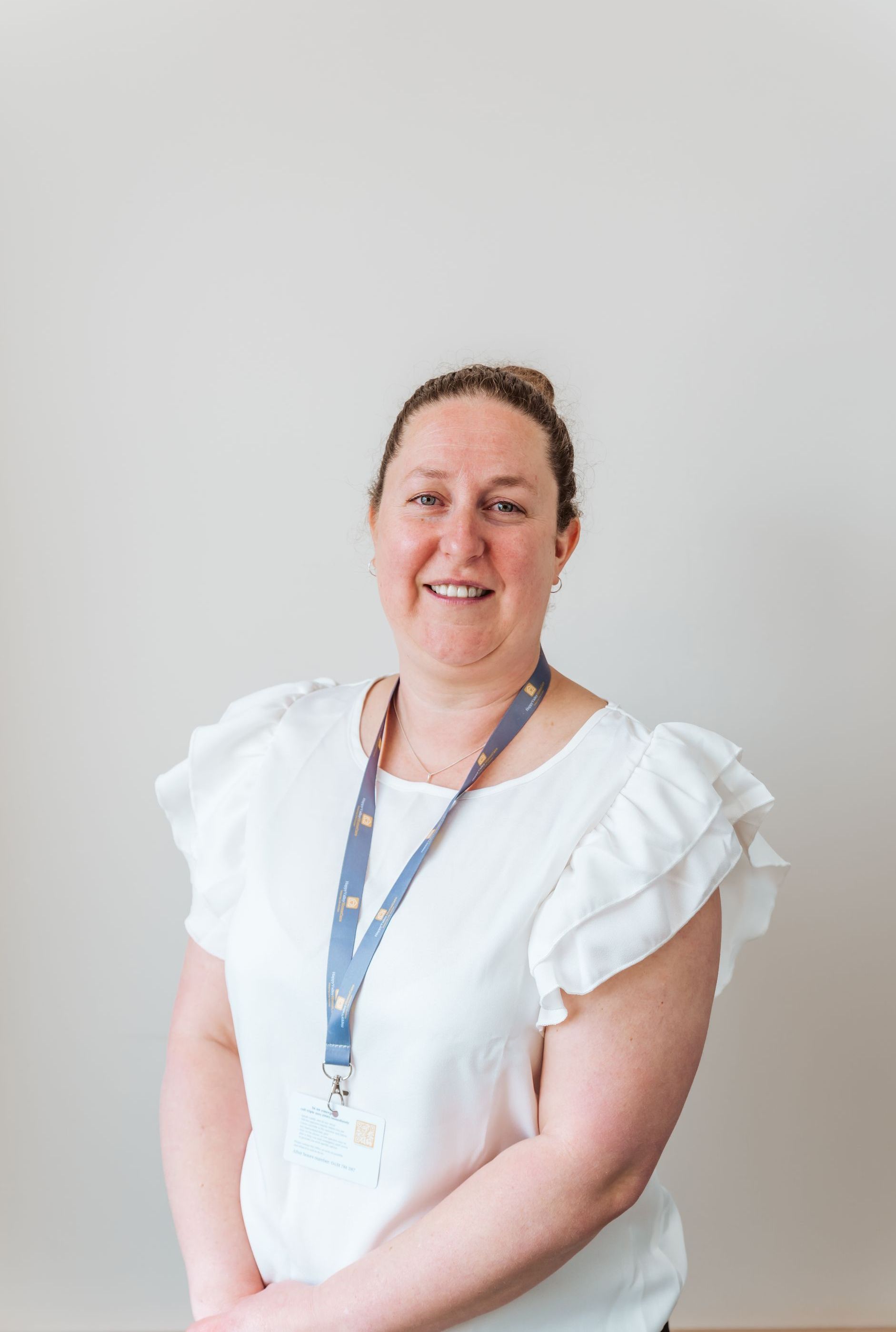 Woman in a white blouse and lanyard standing against a plain light wall, smiling at the camera.