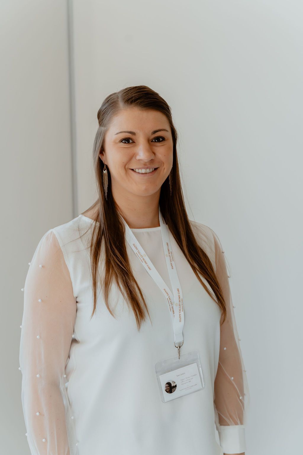 Woman in white blouse and lanyard standing against a plain light wall, smiling at the camera.