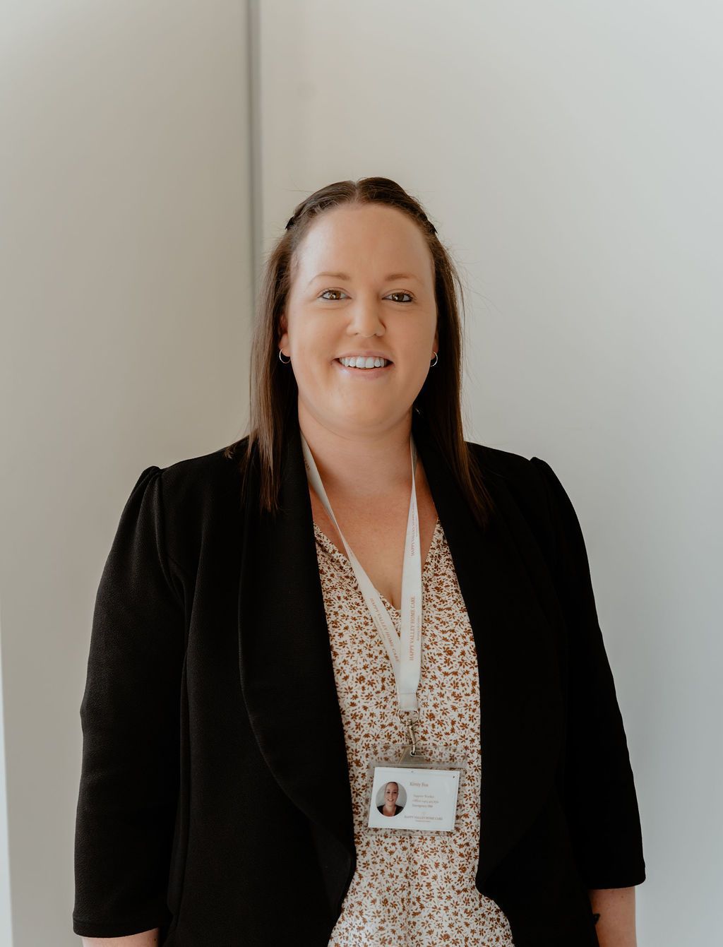 Woman standing indoors against a light wall, wearing a black blazer, patterned blouse, and lanyard badge.