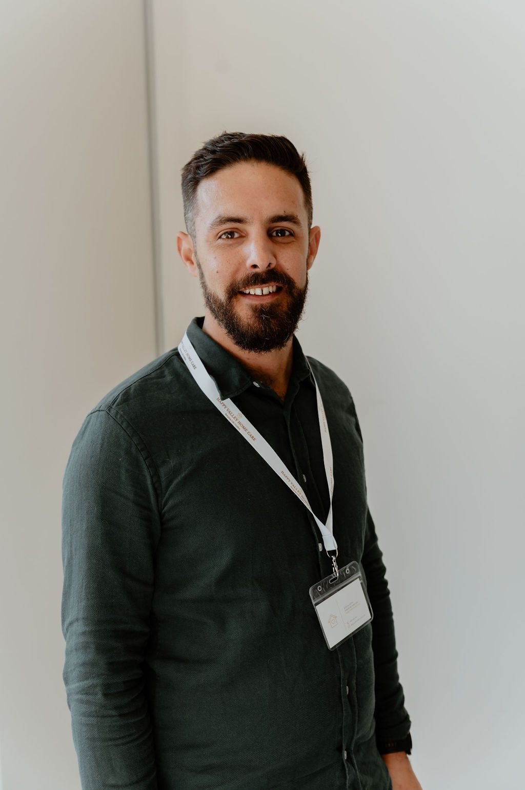 Man standing indoors against a white wall, wearing a dark green shirt and lanyard, looking at the camera