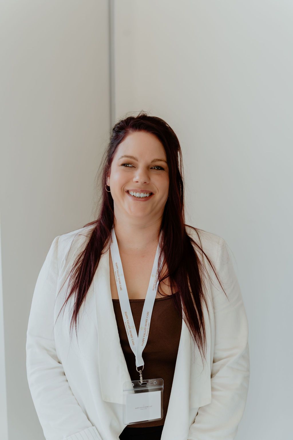 Smiling woman in a white blazer with a lanyard, standing against a plain light wall.