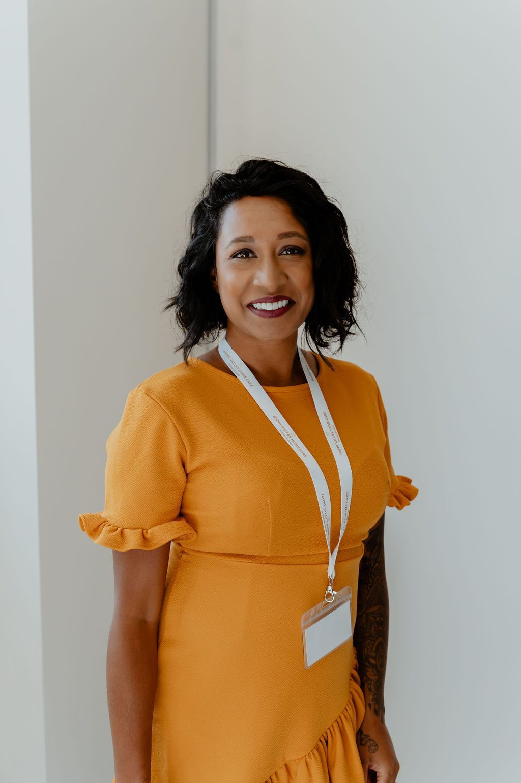 Woman in an orange dress with a lanyard, smiling and standing indoors against a light wall.