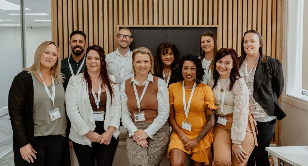 Group of people posing indoors at a conference, wearing lanyards and smiling in front of wood paneling.