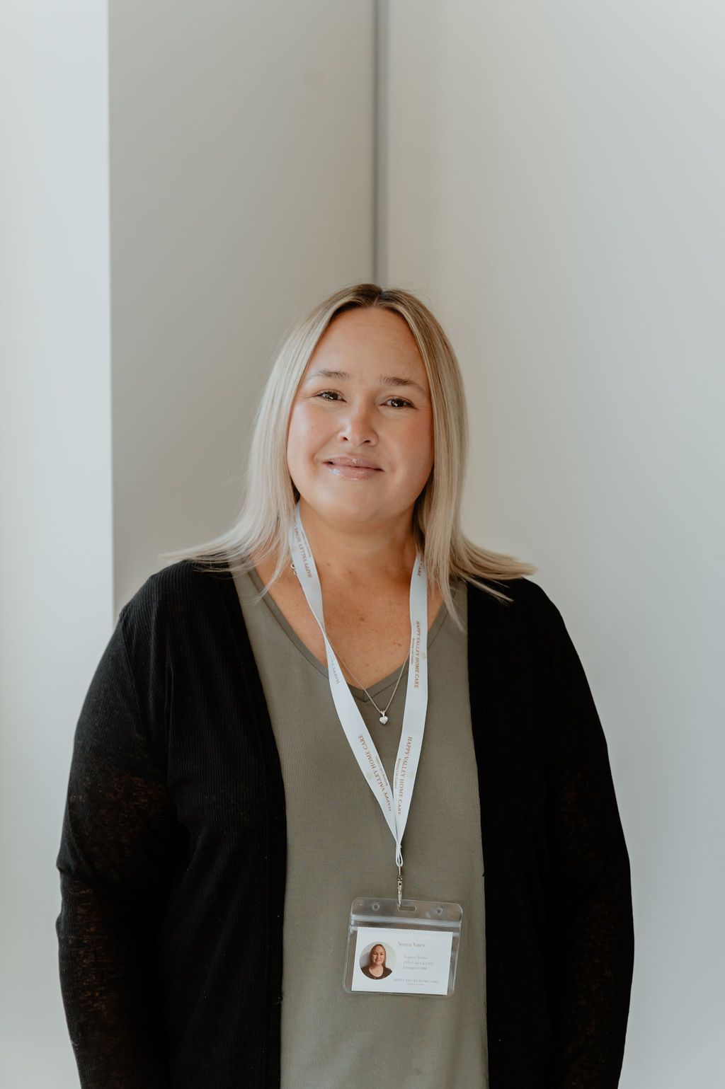 Woman wearing a black cardigan and lanyard, standing against a light wall and smiling gently