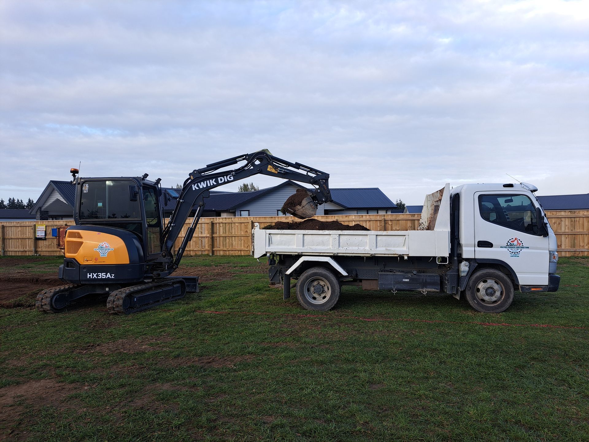 Excavator loading soil into a white dump truck in a grassy construction site