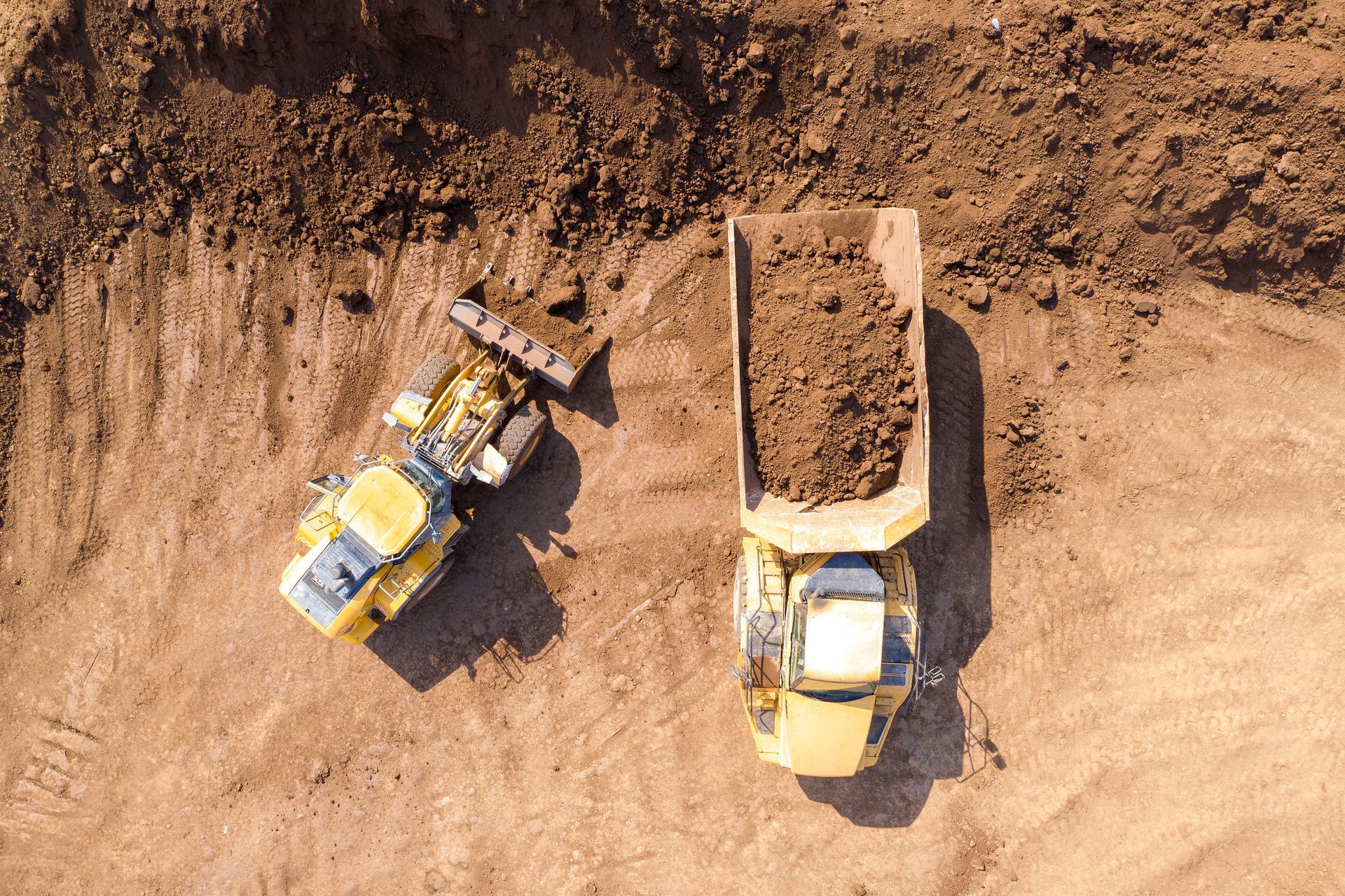 A yellow excavator loading brown soil into a dump truck, both on a brown construction site. A yellow excavator loading brown soil into a dump truck, both on a brown construction site.
