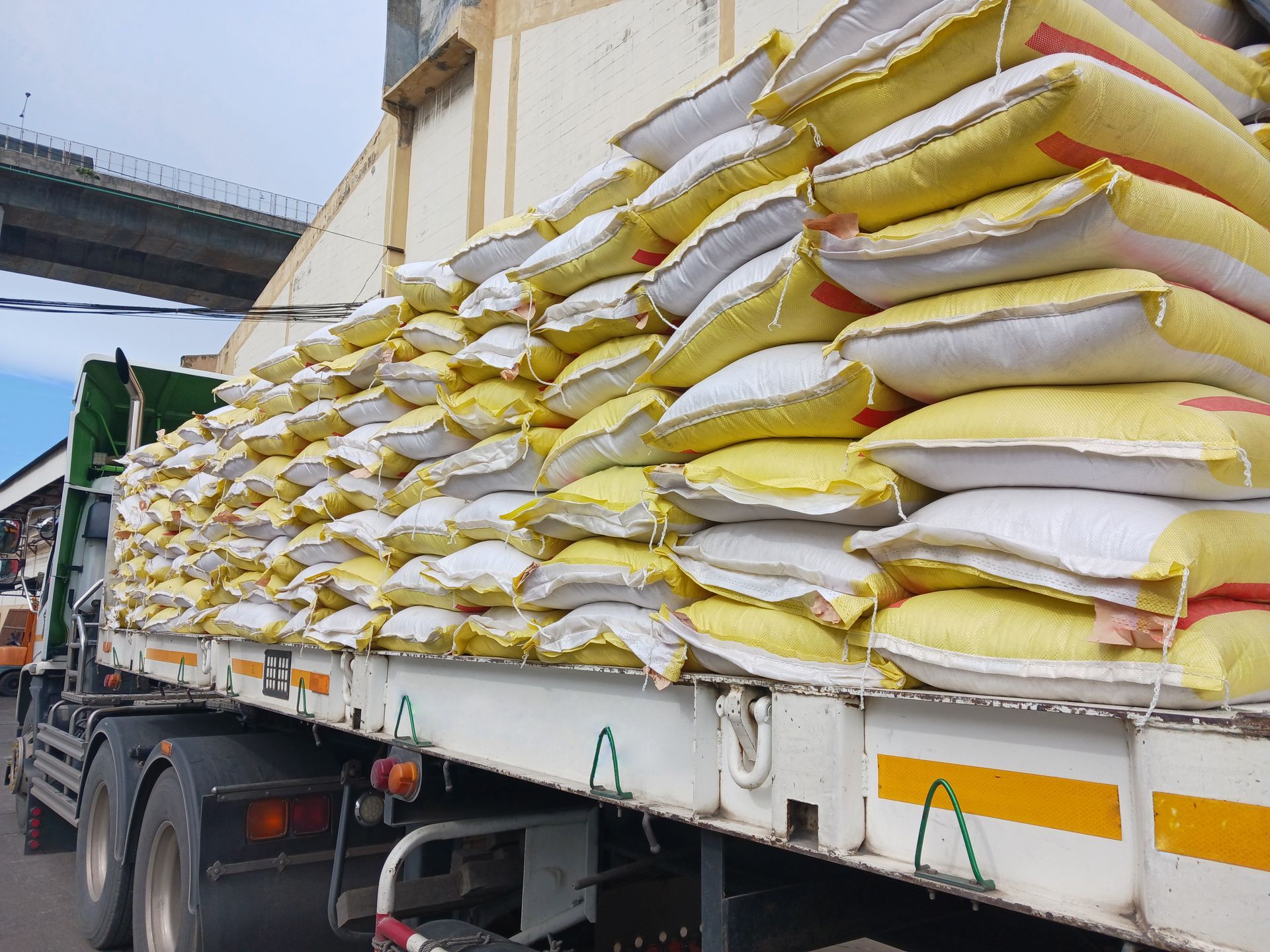 Truck loaded with stacked yellow and white sacks; bridge in background.