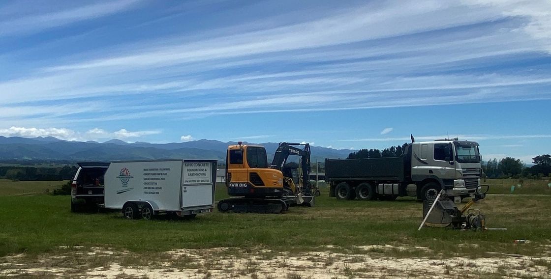 A yellow excavator, a white trailer, and a grey tipper truck parked in a grassy field under a bright blue sky. A yellow excavator, a white trailer, and a grey dump truck parked in a grassy field under a bright blue sky.