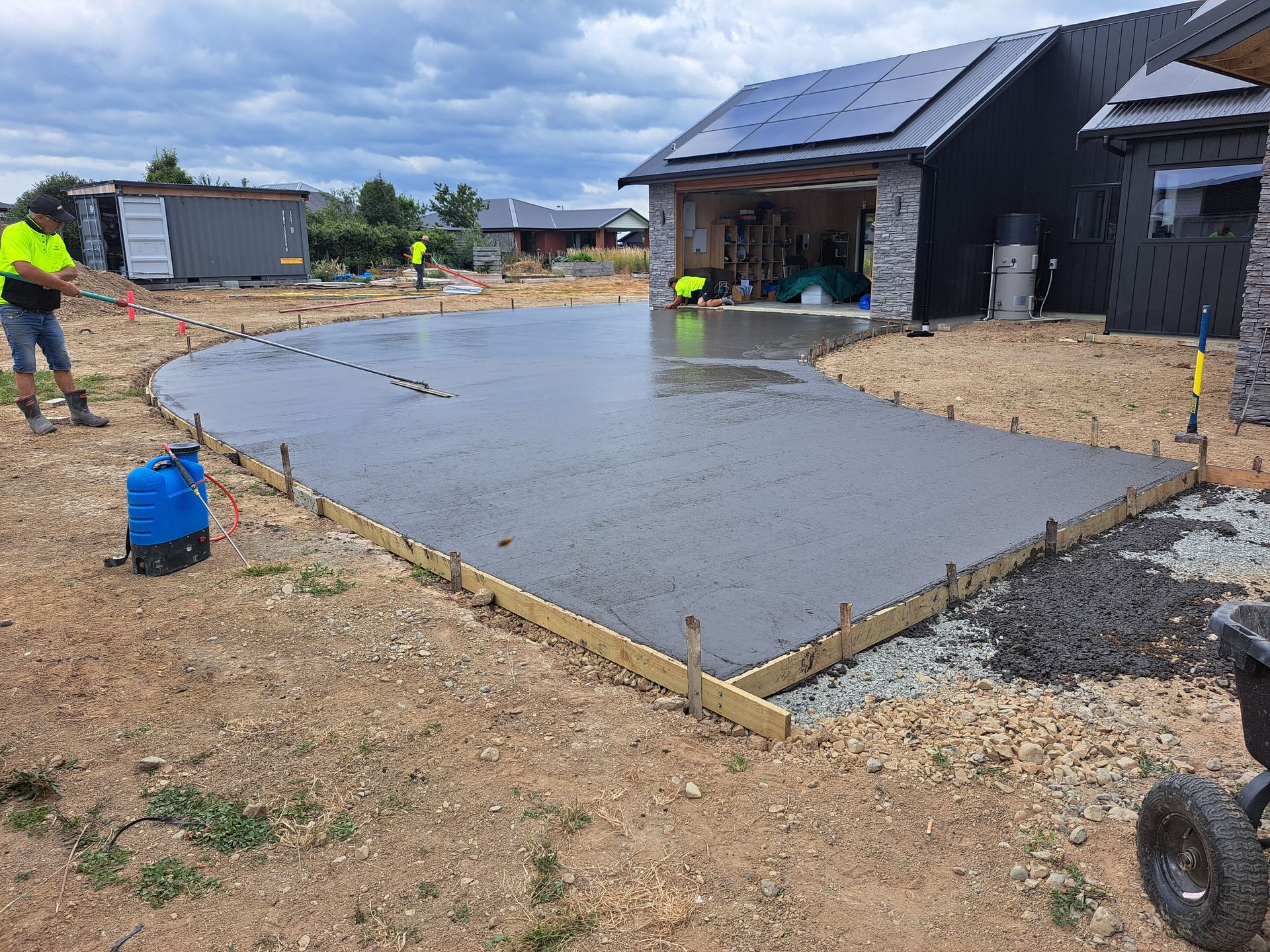 Construction workers finish a fresh concrete driveway slab in front of a modern home with solar panels.