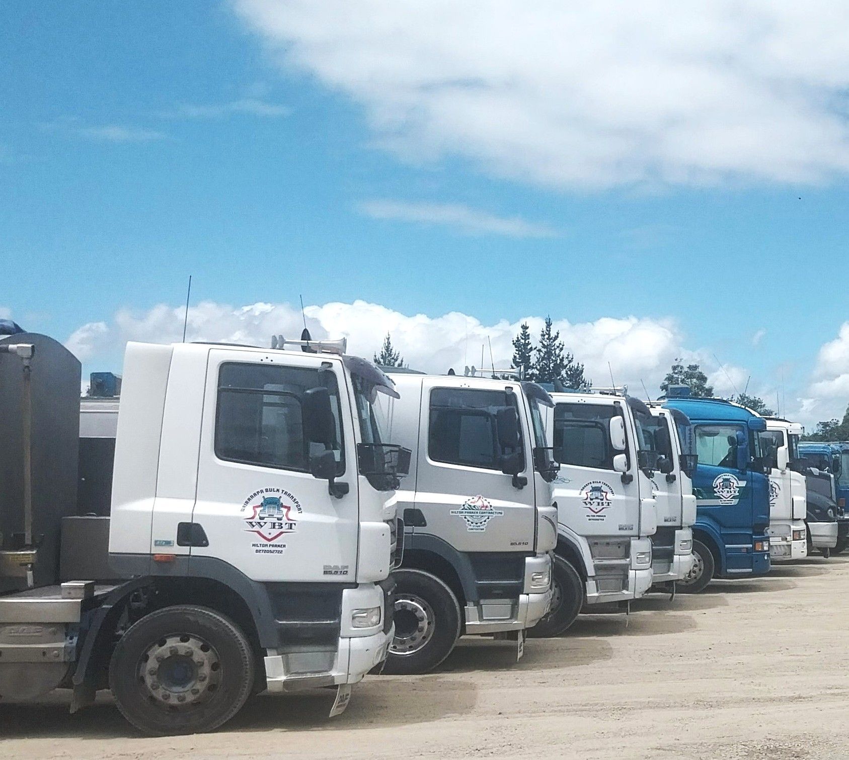 A row of white and blue commercial semi-trucks parked in a gravel lot under a bright, cloudy blue sky.