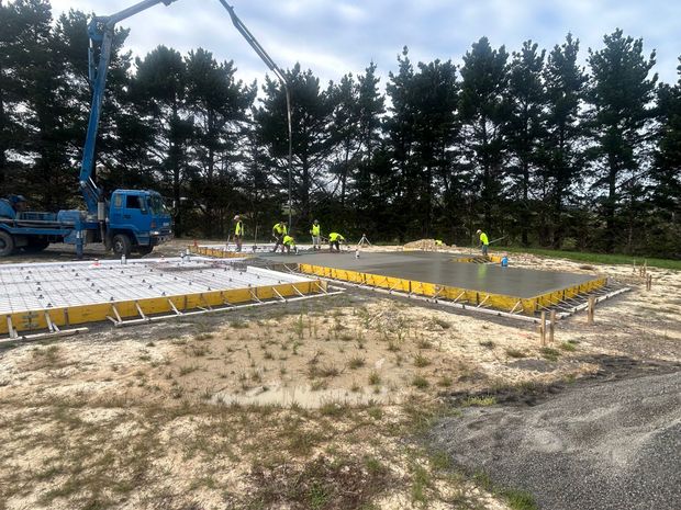A blue concrete pump truck pours cement into a yellow-framed foundation at a construction site with trees in the background. A blue concrete pump truck pours cement into a yellow-framed foundation at a construction site with trees in the background.