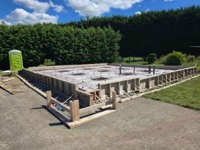 A concrete foundation construction site with wooden formwork, steel rebar reinforcement, and plumbing pipes visible.