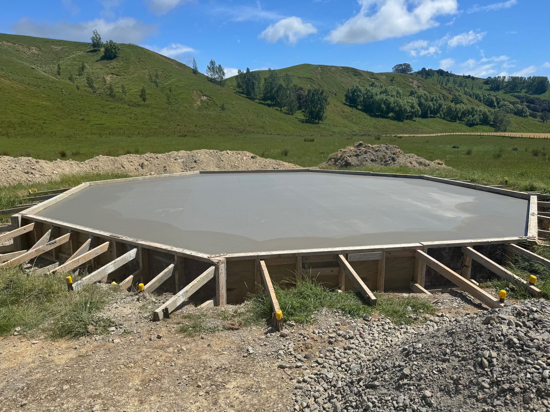 A fresh, octagonal concrete foundation slab poured within wooden formwork on a grassy, hilly construction site. A fresh, octagonal concrete foundation slab poured within wooden formwork on a grassy, hilly construction site.