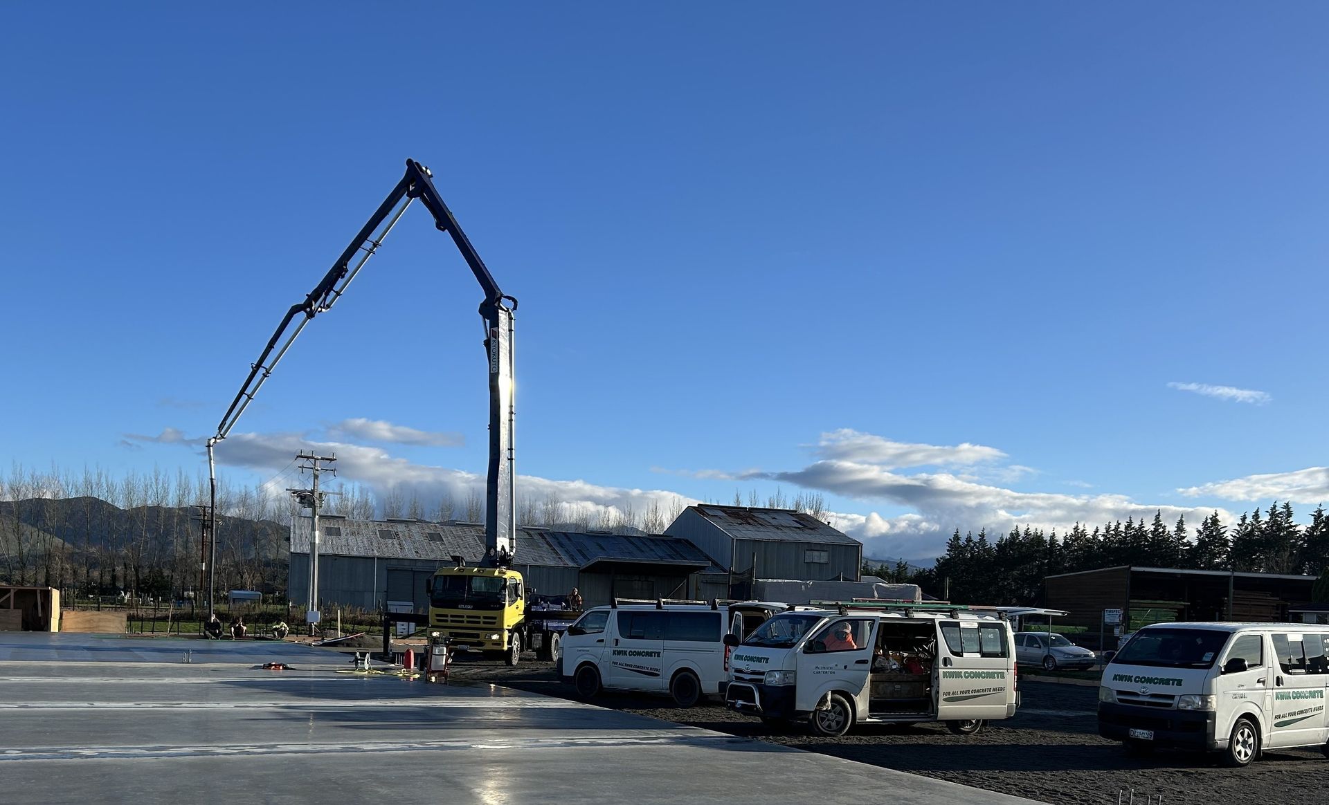 A yellow concrete pump truck extending its arm over a construction site with several white utility vans parked nearby. A yellow concrete pump truck extending its arm over a construction site with several white utility vans parked nearby.