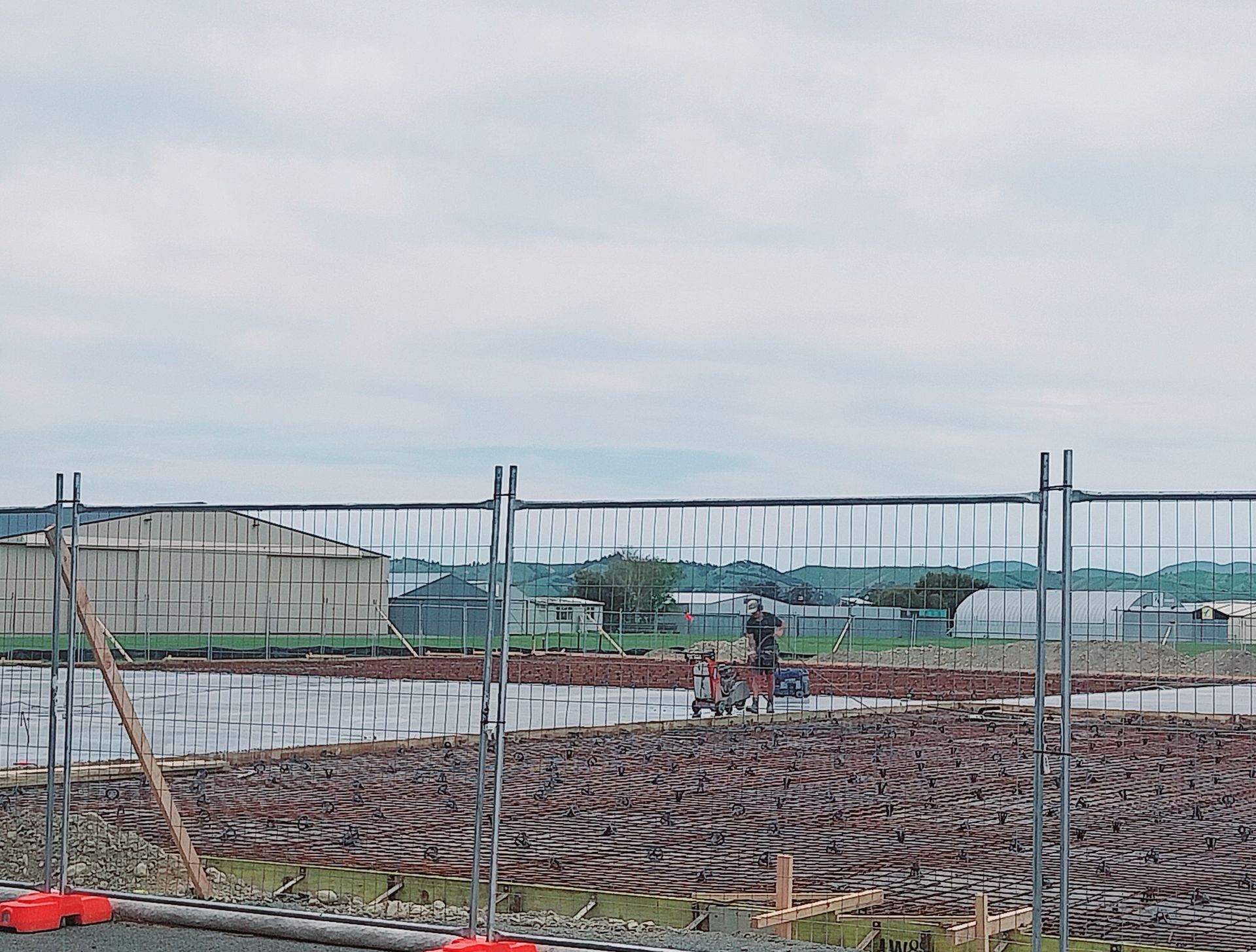 A person stands in a muddy, tilled field behind a chain-link fence, with industrial buildings visible in the distance. A person stands in a muddy, tilled field behind a chain-link fence, with industrial buildings visible in the distance.