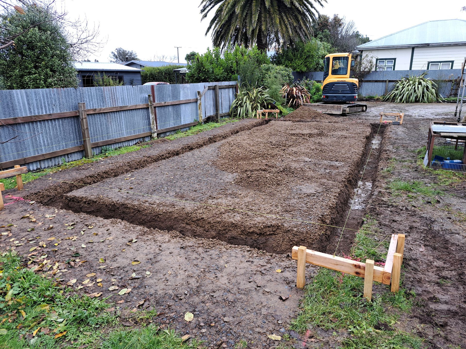 A rectangular trench excavated in a muddy backyard, bordered by wooden formwork and a yellow excavator in the background. A rectangular trench excavated in a muddy backyard, bordered by wooden formwork and a yellow excavator in the background.