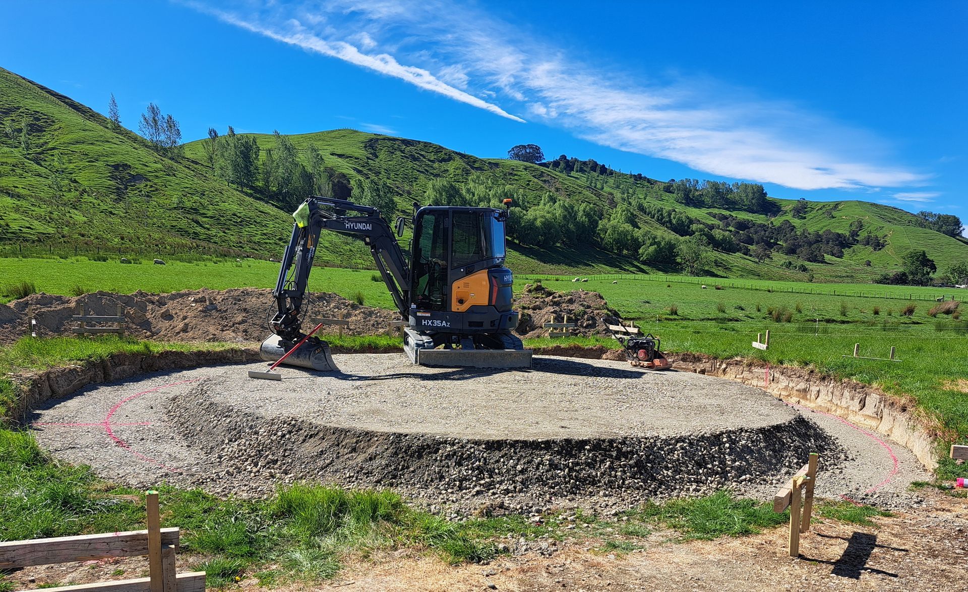 A small, black and yellow excavator sits atop a raised, circular gravel pad in a grassy field beneath a blue sky. A small, black and yellow excavator sits atop a raised, circular gravel pad in a grassy field beneath a blue sky.