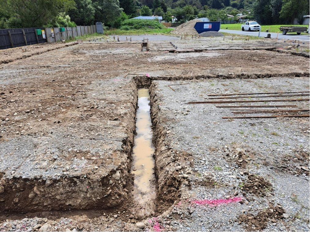 A muddy trench dug into a gravel construction site, with steel reinforcement bars lying on the ground nearby. A muddy trench dug into a gravel construction site, with steel reinforcement bars lying on the ground nearby.