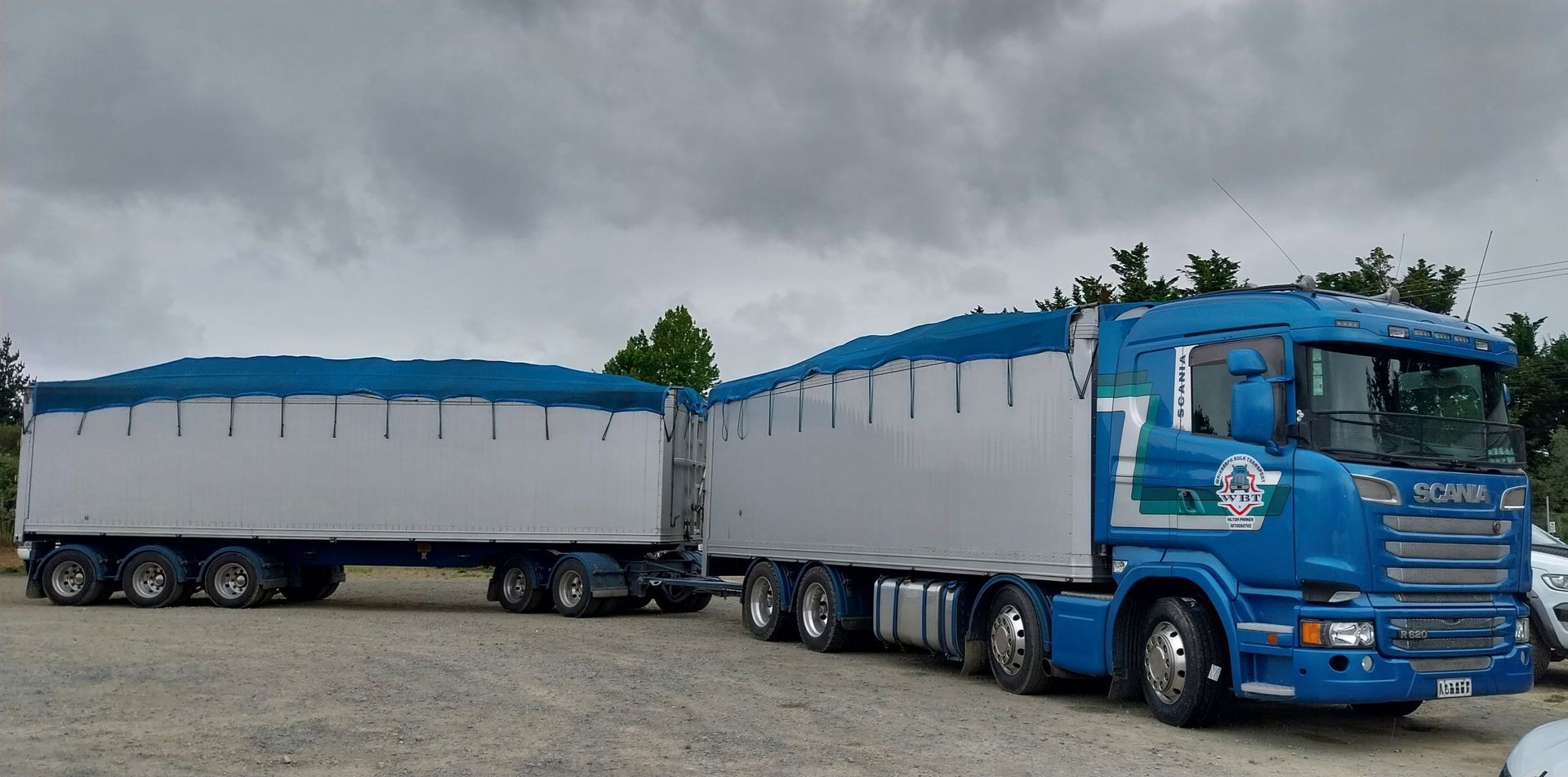 Blue dump truck unloading soil onto grassy area next to a road, house and fence. A blue Scania semi-truck with a silver and blue trailer parked on a gravel lot under a cloudy sky.