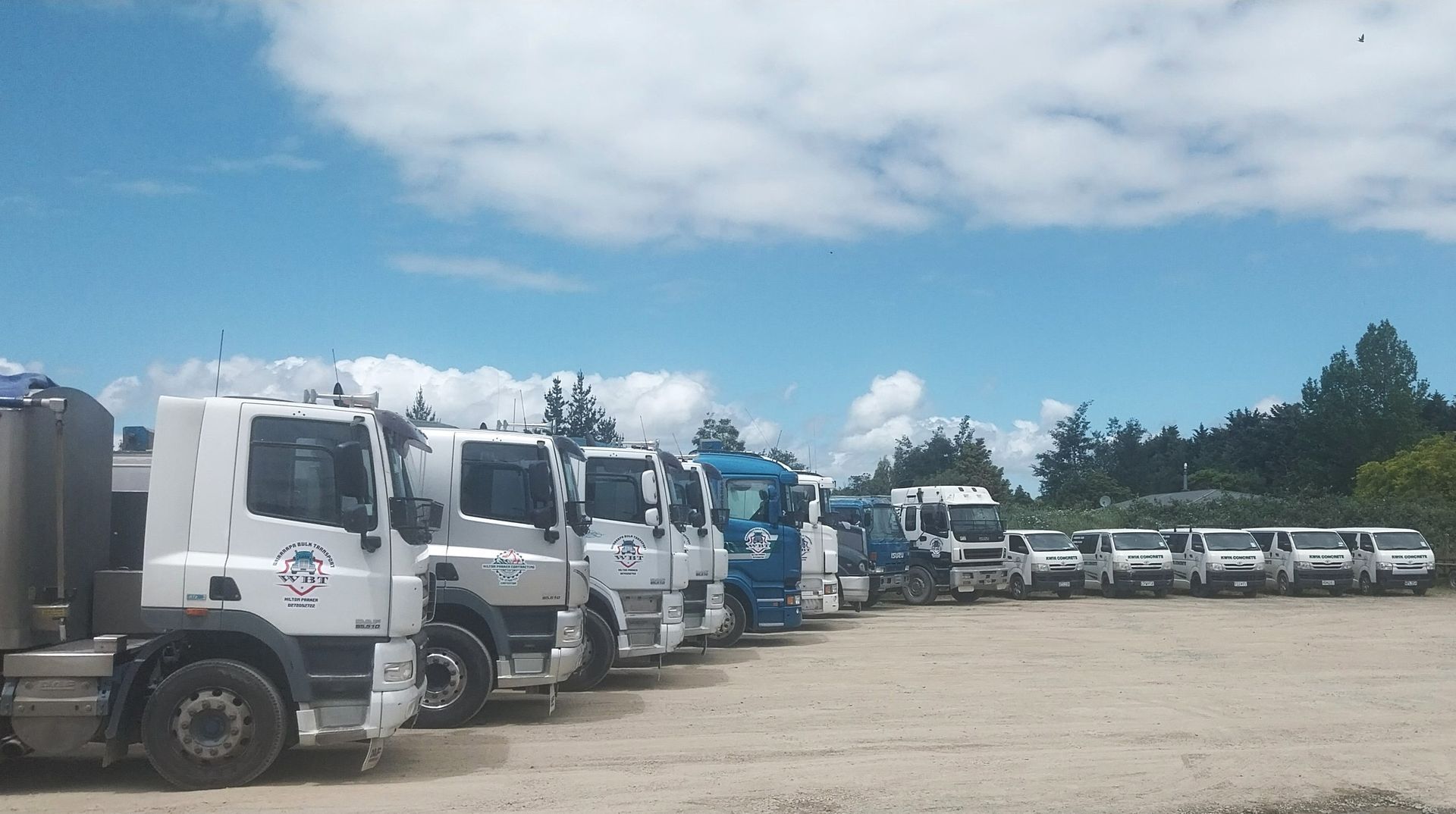 A line of white and blue semi-trucks parked on a gravel lot under a clear sky. A line of white and blue semi-trucks parked on a gravel lot under a clear sky.