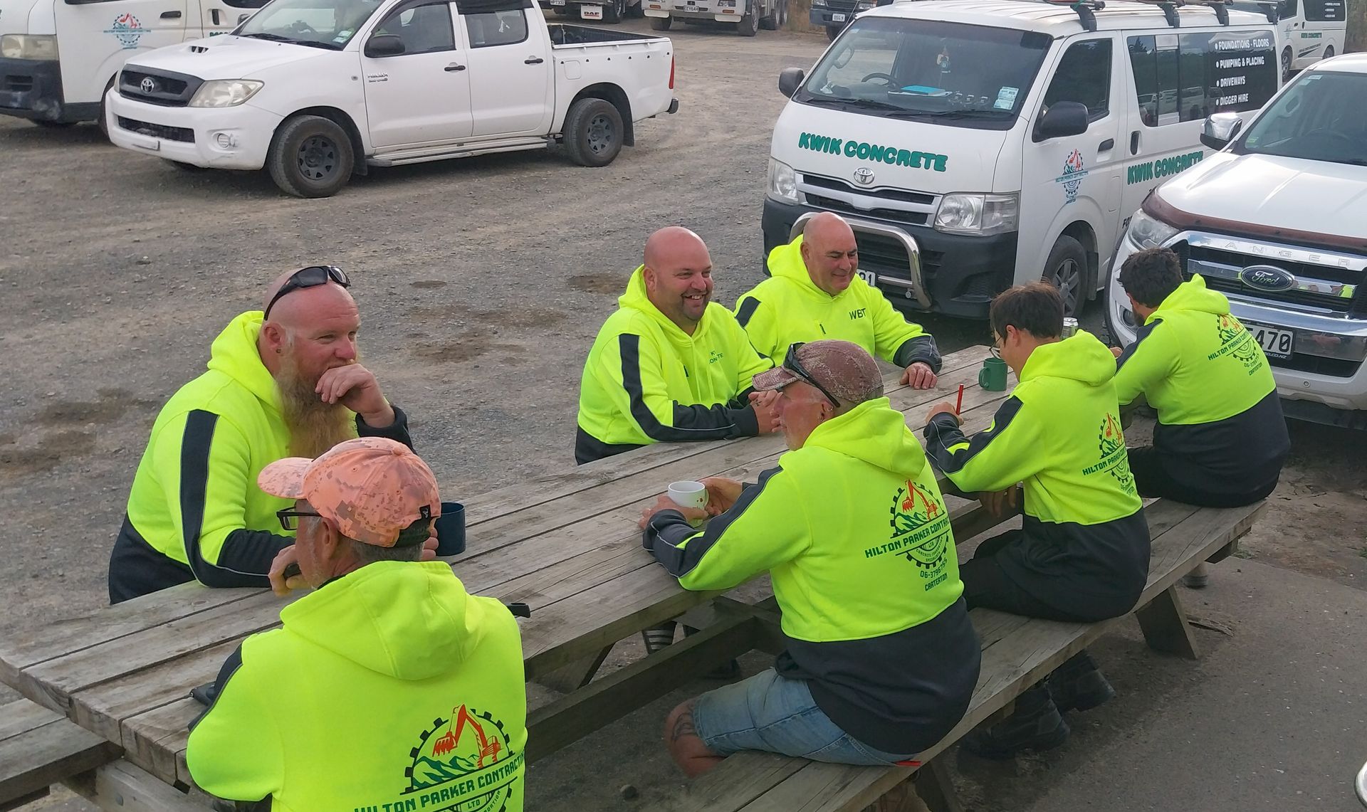 A group of workers in bright yellow high-visibility hoodies sit at an outdoor picnic table near parked service vehicles.