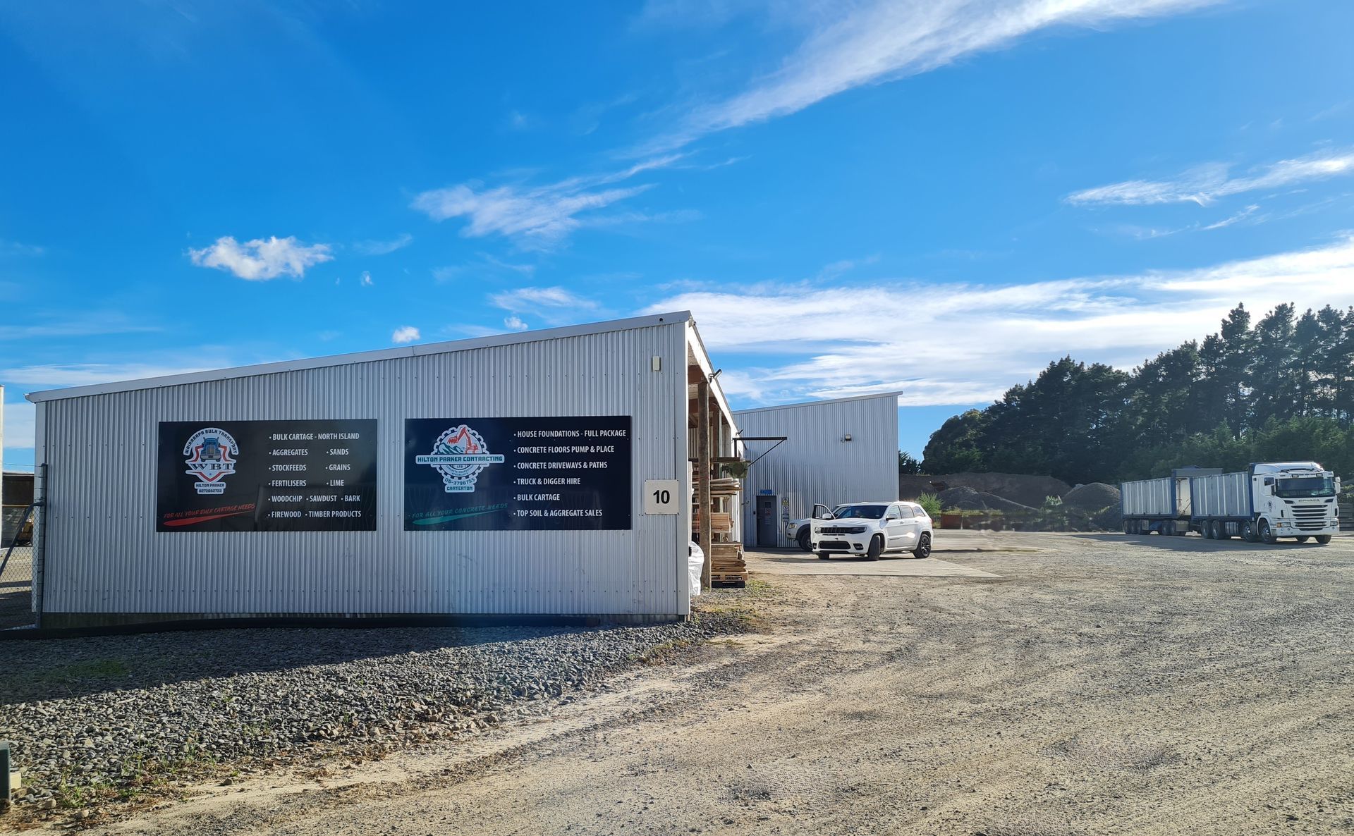 A white shipping container building with business signage on a gravel lot under a bright blue sky, with vehicles nearby.