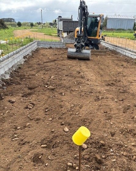 An excavator works inside a concrete foundation wall on a rural construction site, with a yellow marker in the foreground.