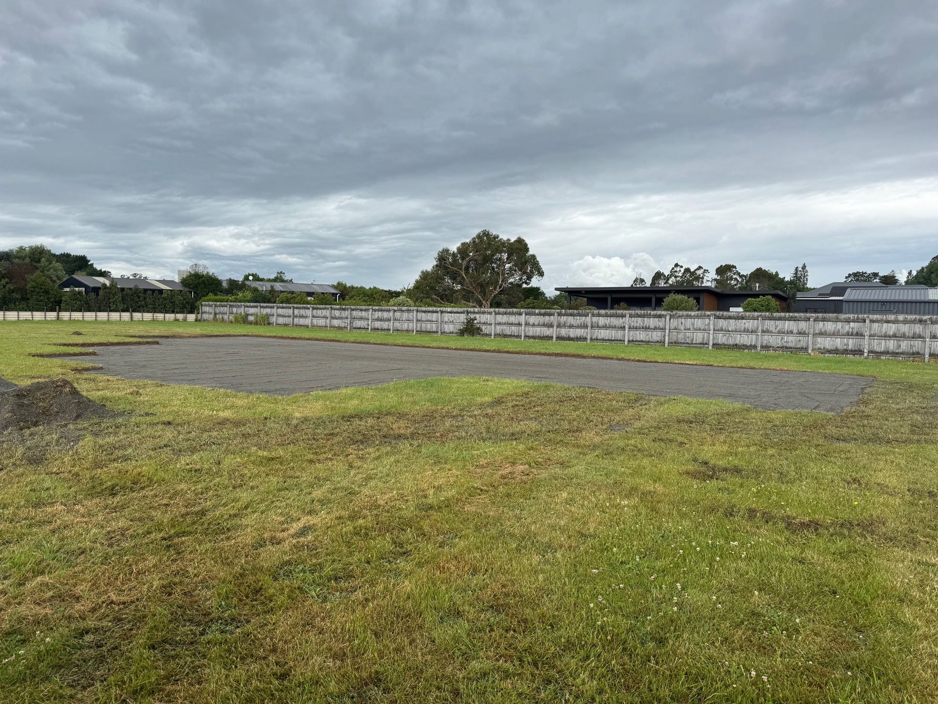 An empty gravel area set within a large grassy field, bordered by a fence under a cloudy sky.