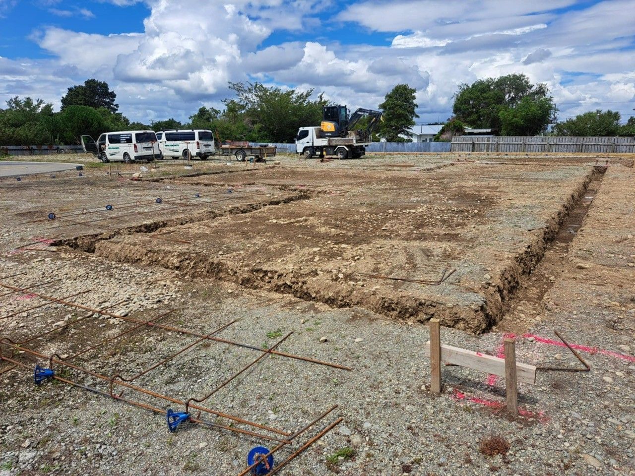 A construction site with excavated foundation trenches for a building, featuring rebar framework and vehicles nearby.