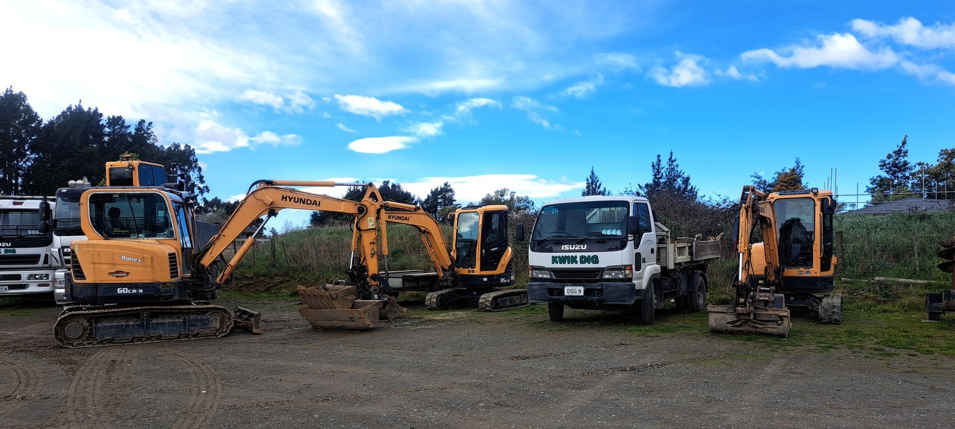 A wide shot of yellow construction excavators and a white dump truck parked on a gravel lot under a bright, sunny sky.