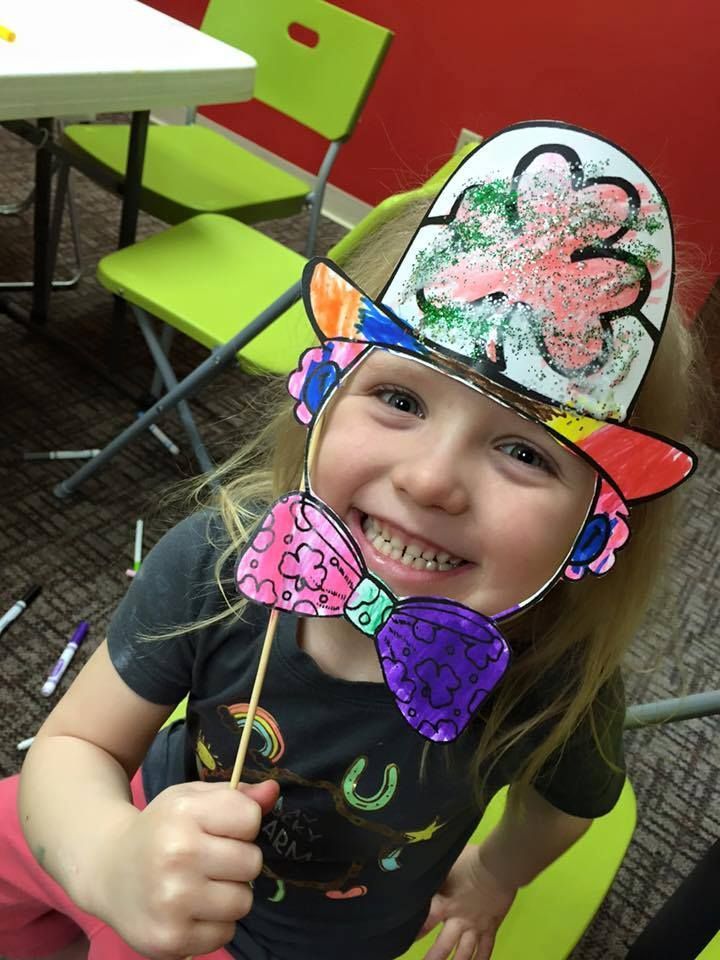 Smiling child wearing a colorful paper hat and shamrock hat at a table in a classroom