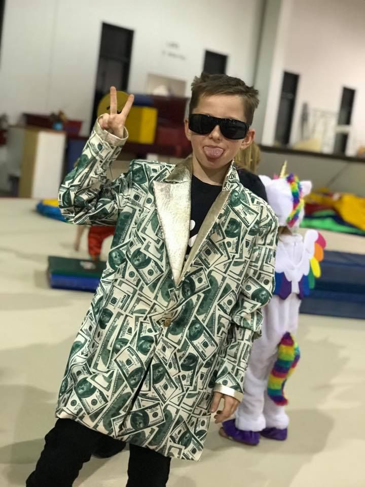 Person in money-print coat and sunglasses poses indoors, flashing a peace sign.
