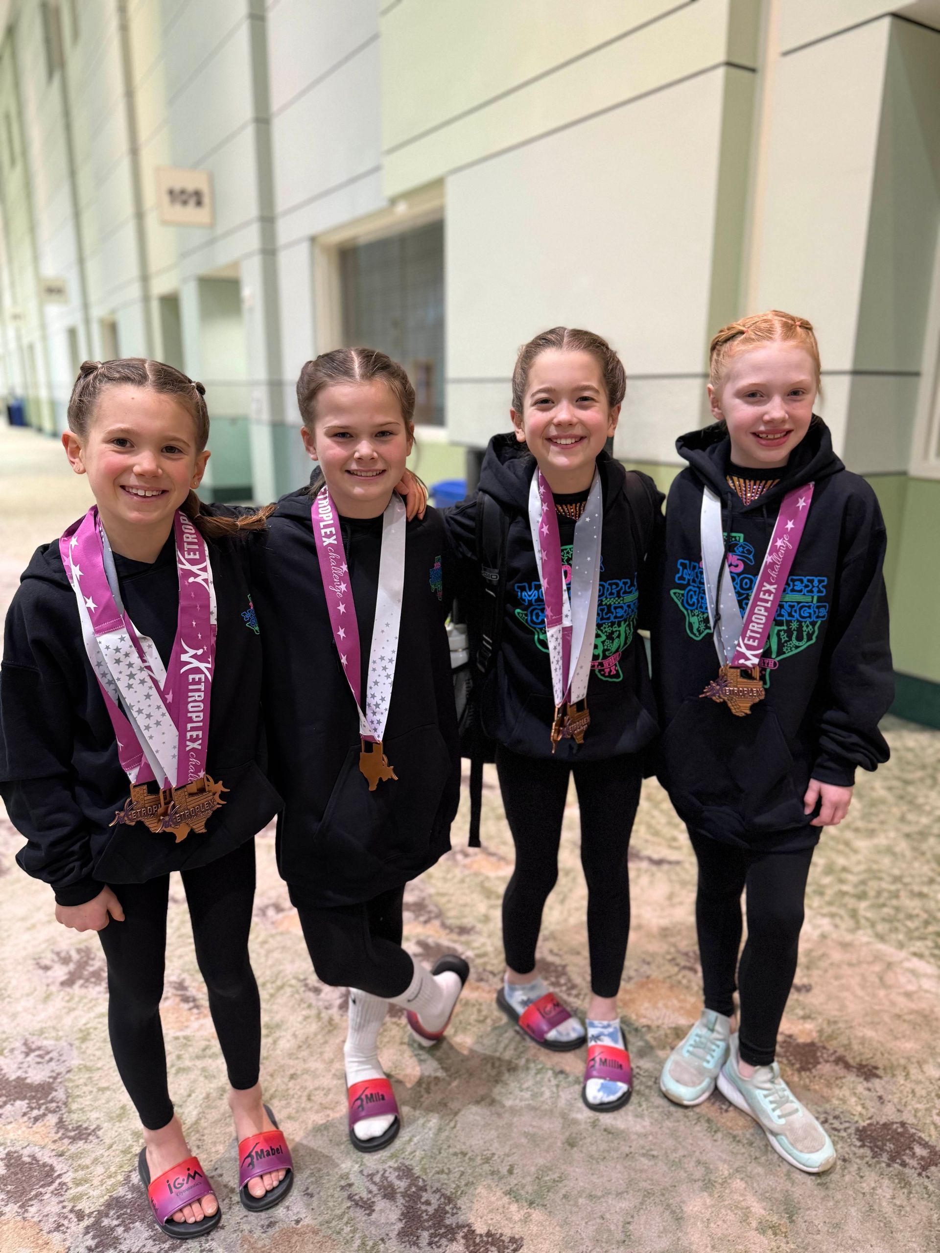 Four girls in black outfits and medals stand smiling indoors after a dance competition.