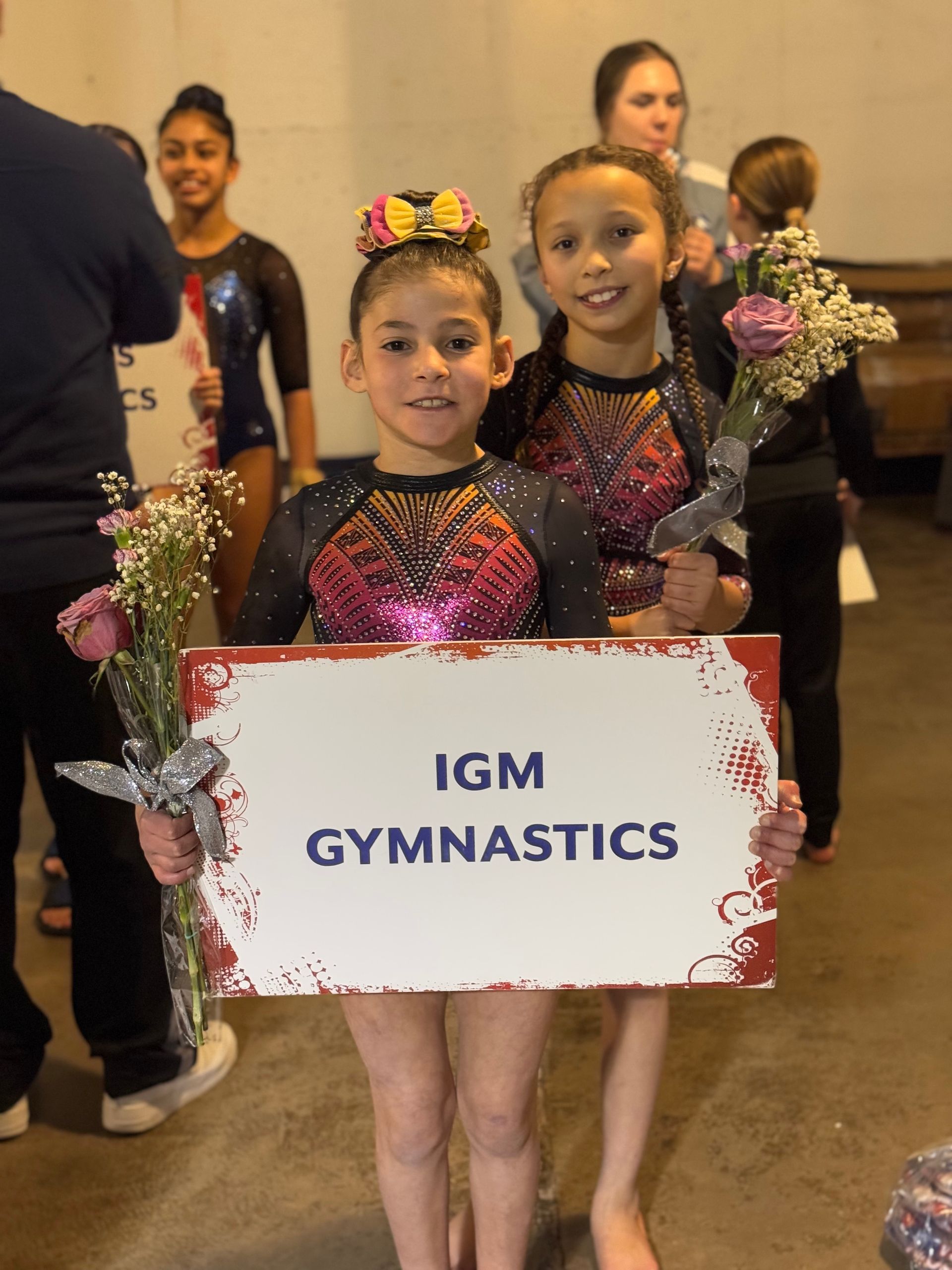 Two gymnasts holding a sign that reads “IGM Gymnastics” with a bouquet in front of a group backstage