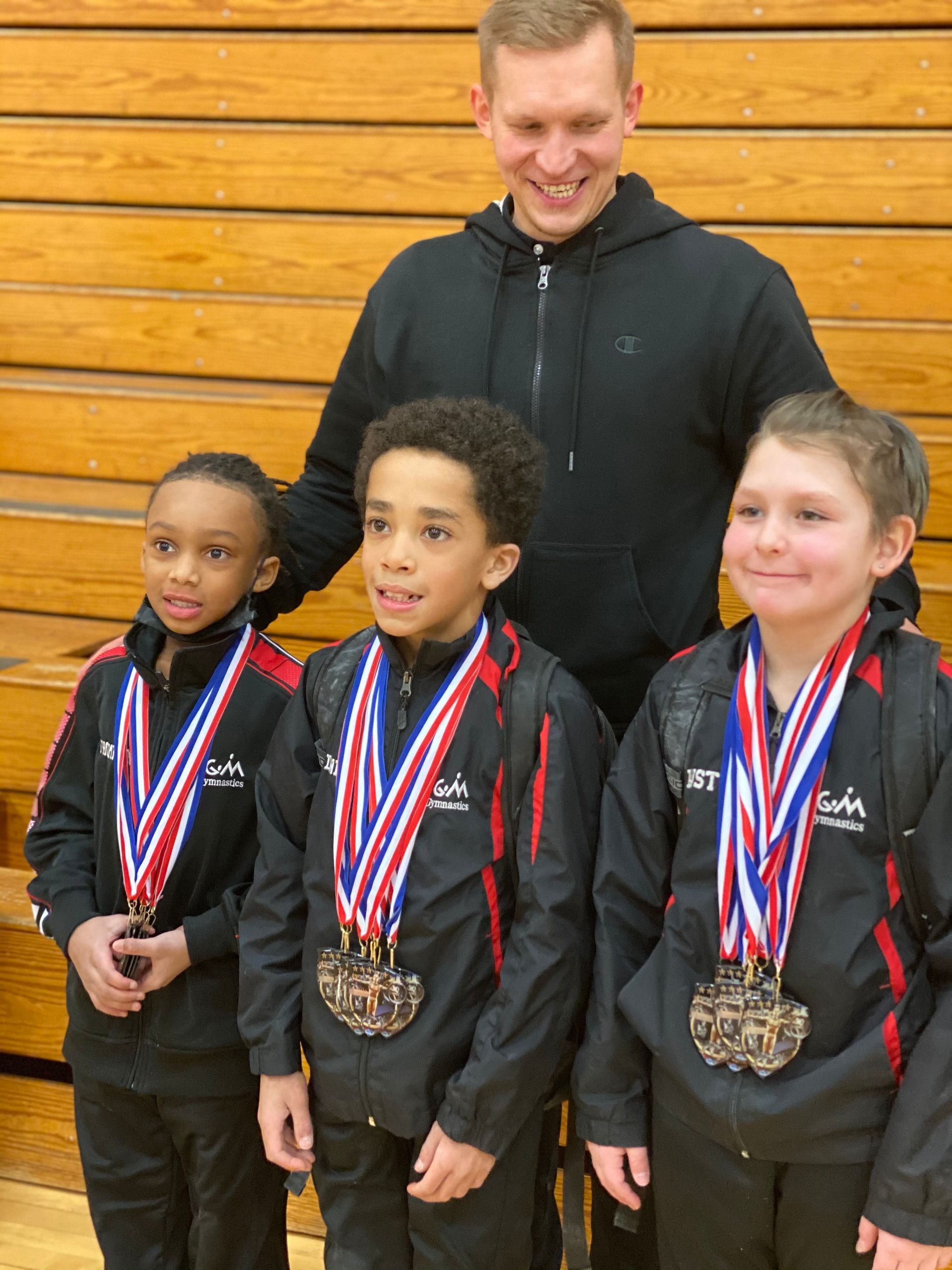 Three medal-winning children in black jackets stand with a smiling coach against a wooden wall.