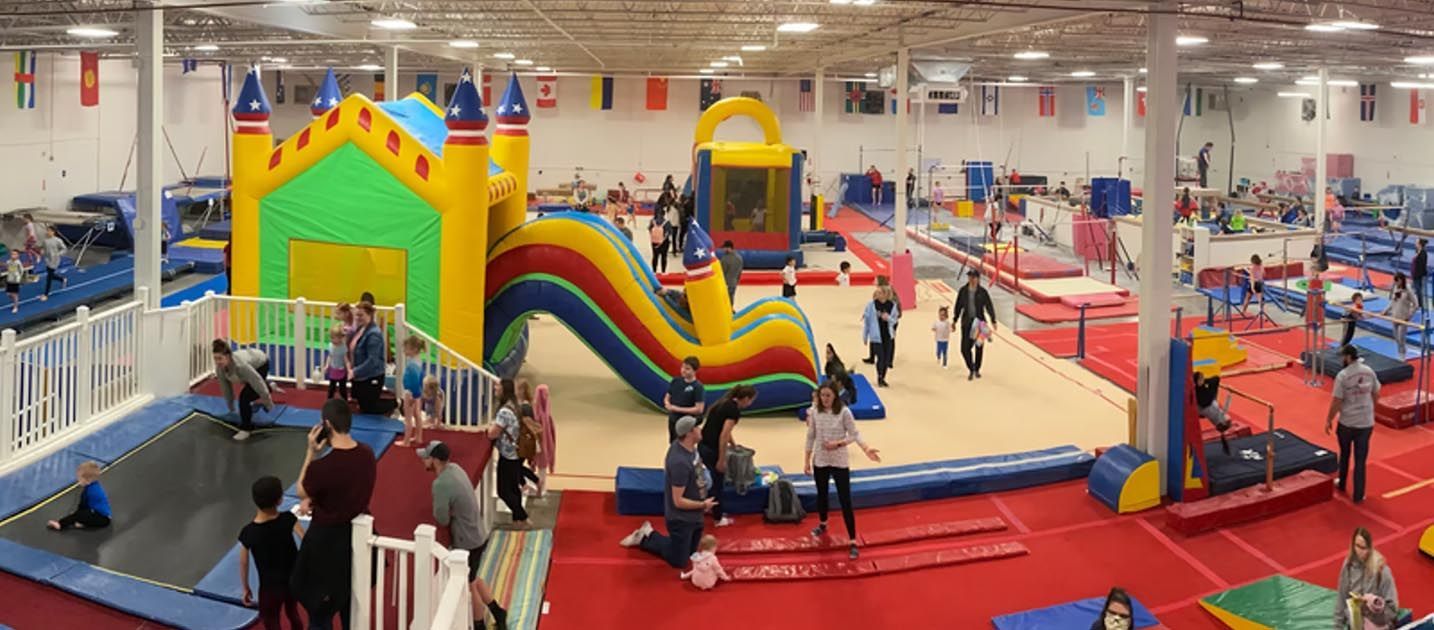 Gymnastics gym with kids and adults. Children on equipment, others watching. Blue, red, and yellow mats.
