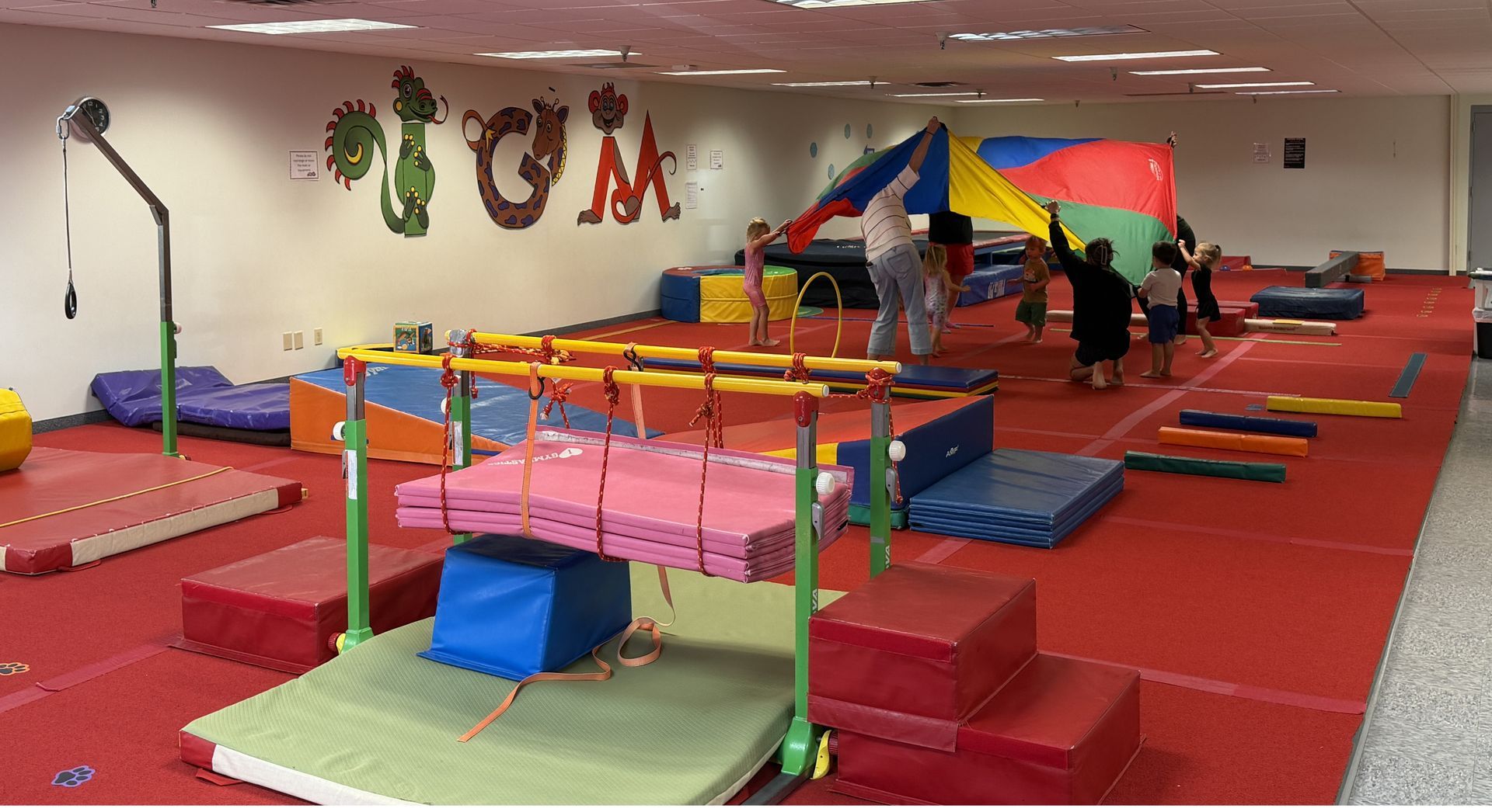 Indoor gymnastics play area with colorful mats, balance bars, and children under a rainbow parachute.
