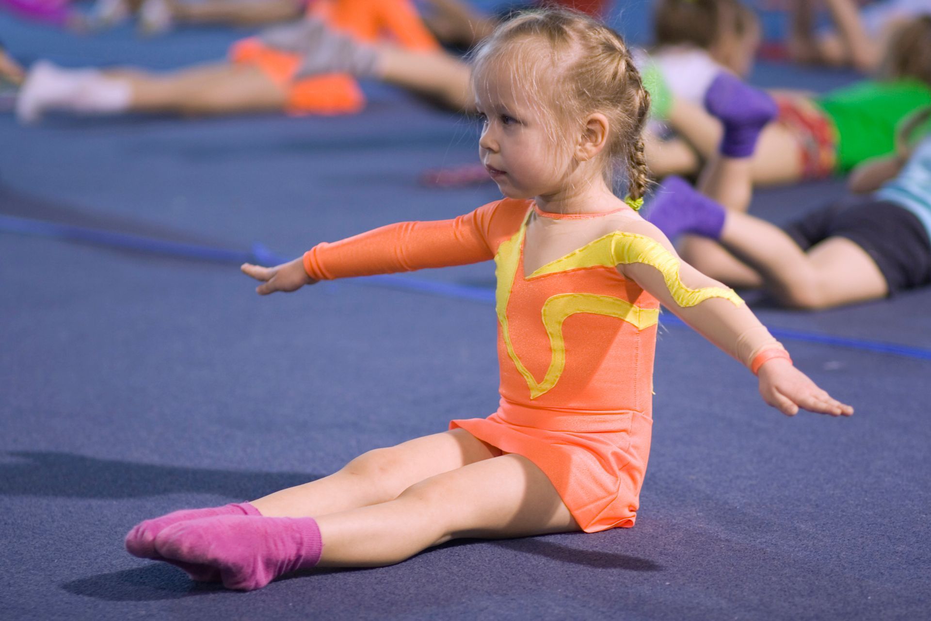 Child doing a somersault with help from an adult on a blue mat in a gym.