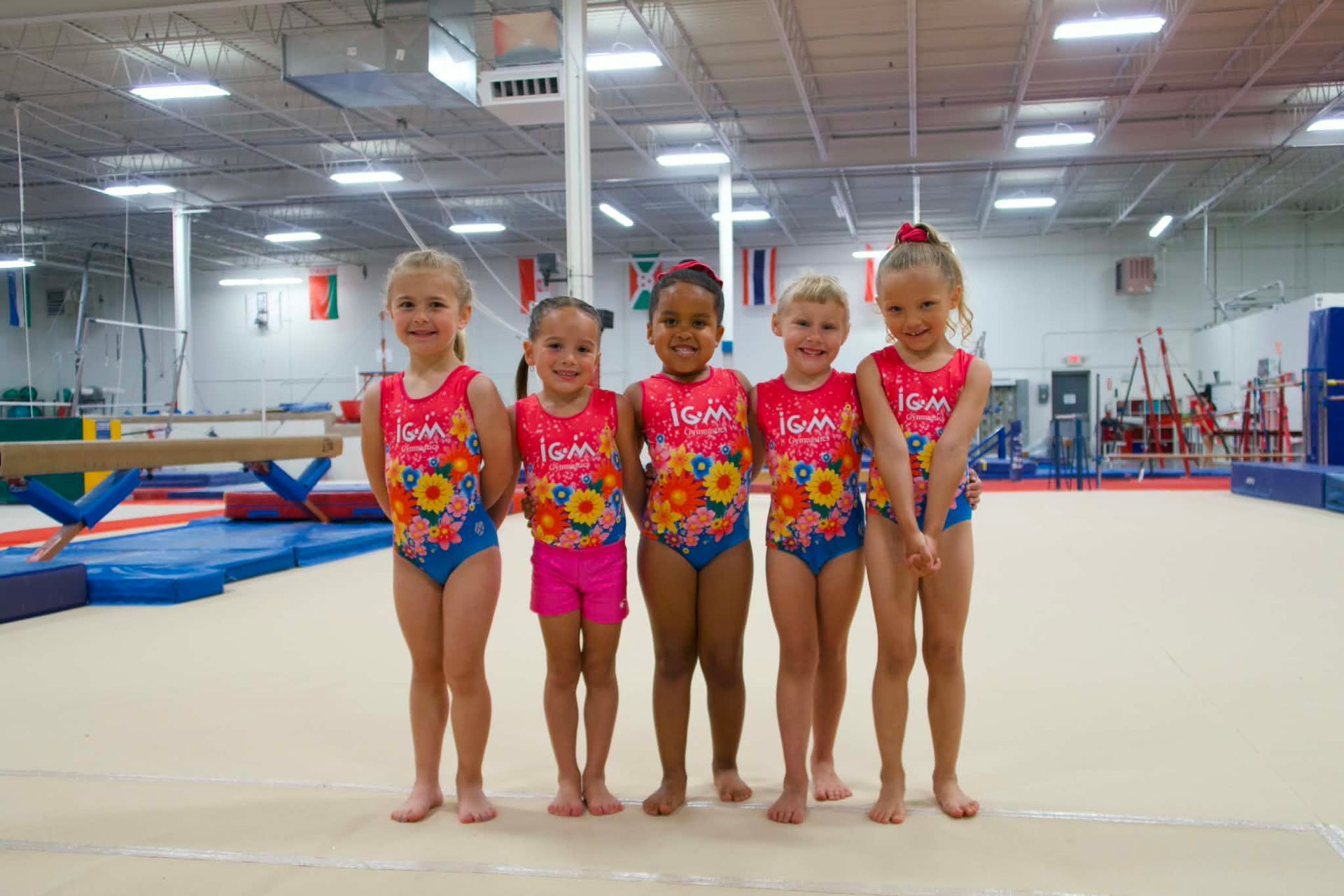 Five gymnasts in pink leotards standing on a gymnastics floor in an indoor training gym