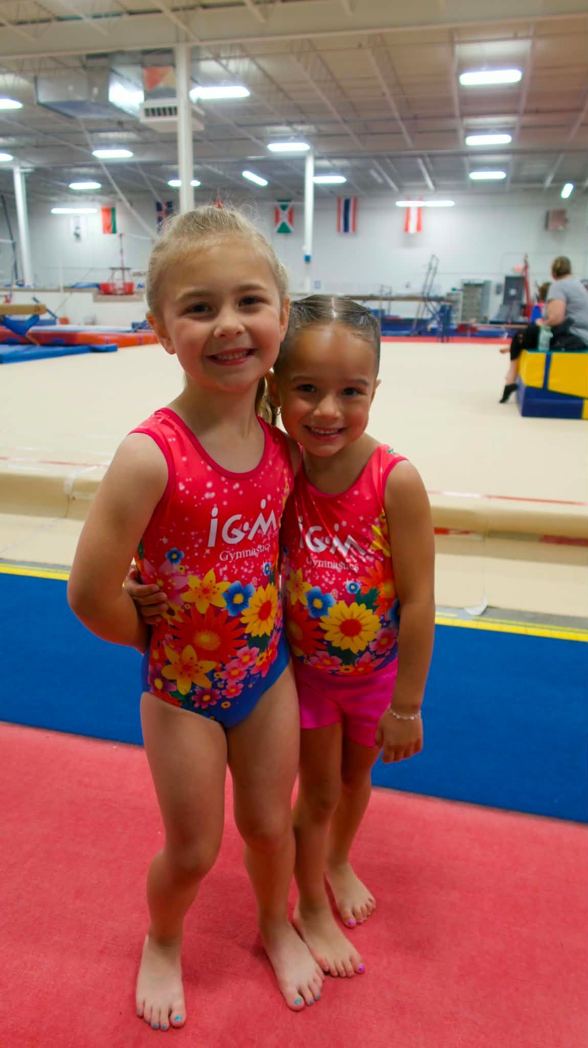 Two children in colorful leotards smiling on a gymnastics mat in a gym